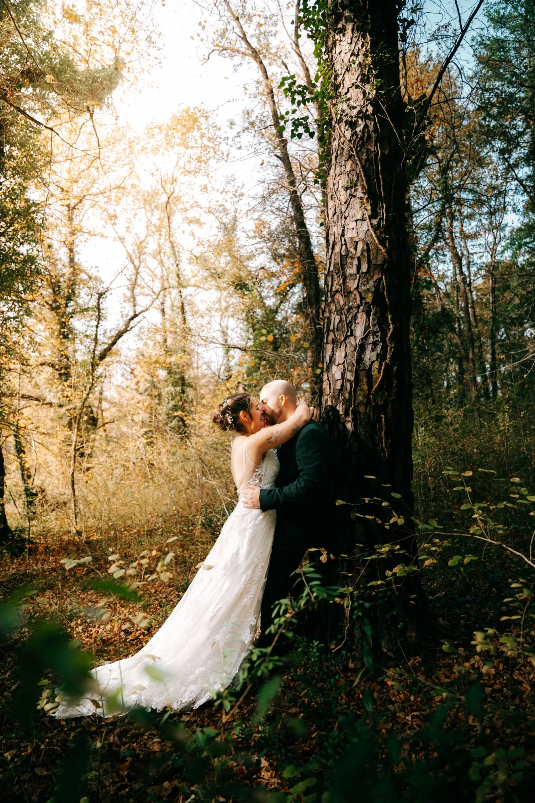 Los novios se besan apoyados en un gran árbol, con la luz dorada del bosque